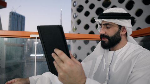 تعابير السرور والسعادة، استخدام جهاز تقني حديث ومتطور، تصوير مقرب لرجل عربي خليجي اماراتي يرتدي الكندورة والغترة البيضاء يستخدم الجهاز اللوحي، إنجاز مهام العمل عن بعد، إدارة شؤون الأعمال وتنظيمها عبر الايباد