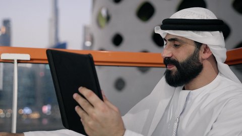 ايماءات السرور والسعادة، استخدام جهاز تقني حديث ومتطور، تصوير مقرب لرجل عربي خليجي اماراتي يرتدي الكندورة والغترة البيضاء يستخدم الجهاز اللوحي، إنجاز مهام العمل عن بعد، إدارة شؤون الأعمال وتنظيمها عبر الايباد