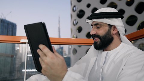 تعابير السرور والسعادة، استخدام جهاز تقني حديث ومتطور، تصوير مقرب لرجل عربي خليجي اماراتي يرتدي الكندورة والغترة البيضاء يستخدم الجهاز اللوحي، إنجاز مهام العمل عن بعد، إدارة شؤون الأعمال وتنظيمها عبر الايباد