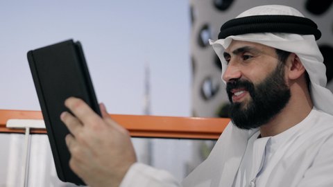 ايماءات السرور والسعادة، استخدام جهاز تقني حديث ومتطور، تصوير مقرب لرجل عربي خليجي اماراتي يرتدي الكندورة والغترة البيضاء يستخدم الجهاز اللوحي، إنجاز مهام العمل عن بعد، إدارة شؤون الأعمال وتنظيمها عبر الايباد