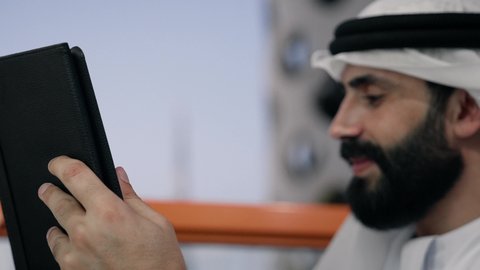 إنجاز مهام العمل عن بعد، إدارة شؤون الأعمال وتنظيمها عبر الايباد، ايماءات السرور والسعادة، استخدام جهاز تقني حديث ومتطور، تصوير مقرب لرجل عربي خليجي اماراتي يرتدي الكندورة والغترة البيضاء يستخدم الجهاز اللوحي