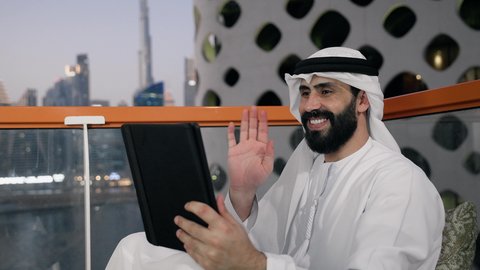 التواصل و إجراء مكالمة فيديو، إنجاز مهام العمل عن بعد، إدارة شؤون الأعمال وتنظيمها عبر الايباد، استخدام جهاز تقني حديث ومتطور، تصوير مقرب لرجل عربي خليجي اماراتي يرتدي الكندورة والغترة البيضاء يستخدم الجهاز اللوحي ، رفع كف اليد بايماءات القاء التحية والسلام