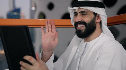 رفع كف اليد بايماءات القاء التحية والسلام، إجراء مكالمة فيديو، إنجاز مهام العمل عن بعد، إدارة شؤون الأعمال وتنظيمها عبر الايباد، استخدام جهاز تقني حديث ومتطور، تصوير مقرب لرجل عربي خليجي اماراتي يرتدي الكندورة والغترة البيضاء يستخدم الجهاز اللوحي