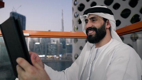 إجراء مكالمة فيديو، إنجاز مهام العمل عن بعد، إدارة شؤون الأعمال وتنظيمها عبر الايباد، استخدام جهاز تقني حديث ومتطور، تصوير مقرب لرجل عربي خليجي اماراتي يرتدي الكندورة والغترة البيضاء يستخدم الجهاز اللوحي ، رفع كف اليد بايماءات القاء التحية والسلام