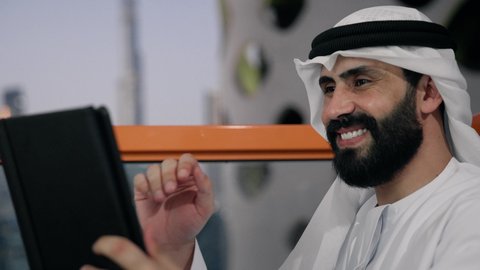 التواصل و إجراء مكالمة فيديو، إنجاز مهام العمل عن بعد، إدارة شؤون الأعمال وتنظيمها عبر الايباد، استخدام جهاز تقني حديث ومتطور، تصوير مقرب لرجل عربي خليجي اماراتي يرتدي الكندورة والغترة البيضاء يستخدم الجهاز اللوحي ، رفع كف اليد بايماءات القاء التحية والسلام