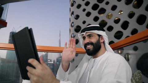 Making a video call, completing work tasks remotely, managing and organizing business affairs via iPad, using a modern and advanced technical device, a close-up of an Emirati Gulf Arab man wearing a white kandura and ghutra using a tablet, raising his palm in greeting and peace gestures
