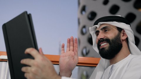 رفع كف اليد بايماءات القاء التحية والسلام، إجراء مكالمة فيديو، إنجاز مهام العمل عن بعد، إدارة شؤون الأعمال وتنظيمها عبر الايباد، استخدام جهاز تقني حديث ومتطور، تصوير مقرب لرجل عربي خليجي اماراتي يرتدي الكندورة والغترة البيضاء يستخدم الجهاز اللوحي