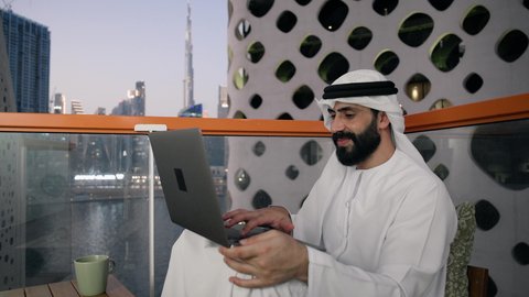إدارة شؤون الأعمال وتنظيمها عبر اللابتوب، ايماءات السعادة والسرور، استخدام جهاز تقني حديث ومتطور، تصوير مقرب لرجل عربي خليجي اماراتي يرتدي الكندورة والغترة البيضاء يستخدم الحاسوب المحمول  ، إنجاز مهام العمل عن بعد