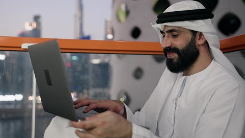 إنجاز مهام العمل عن بعد، إدارة شؤون الأعمال وتنظيمها عبر اللابتوب، ايماءات السعادة والسرور، استخدام جهاز تقني حديث ومتطور، تصوير مقرب لرجل عربي خليجي اماراتي يرتدي الكندورة والغترة البيضاء يستخدم الحاسوب المحمول