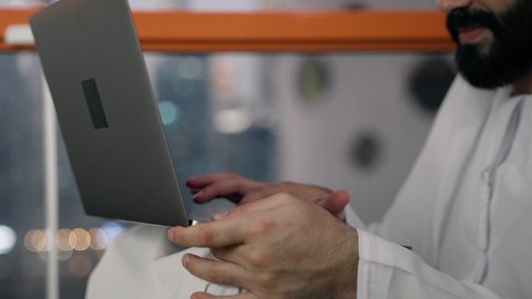إنهاء مهام العمل عن بعد، إدارة شؤون الأعمال وتنظيمها عبر اللابتوب، ايماءات السعادة والسرور، استخدام جهاز تقني حديث ومتطور، تصوير مقرب لرجل عربي خليجي اماراتي يرتدي الكندورة والغترة البيضاء يستخدم الحاسوب المحمول