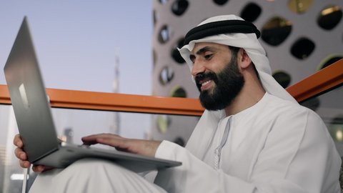 إتمام مهام العمل عن بعد، إدارة شؤون الأعمال وتنظيمها عبر اللابتوب، ايماءات السعادة والسرور، استخدام جهاز تقني حديث ومتطور، تصوير مقرب لرجل عربي خليجي اماراتي يرتدي الكندورة والغترة البيضاء يستخدم الحاسوب المحمول