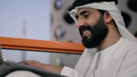 أداء مهام العمل عن بعد، إدارة شؤون الأعمال وتنظيمها عبر اللابتوب، ايماءات السعادة والسرور، استخدام جهاز تقني حديث ومتطور، تصوير مقرب لرجل عربي خليجي اماراتي يرتدي الكندورة والغترة البيضاء يستخدم الحاسوب المحمول