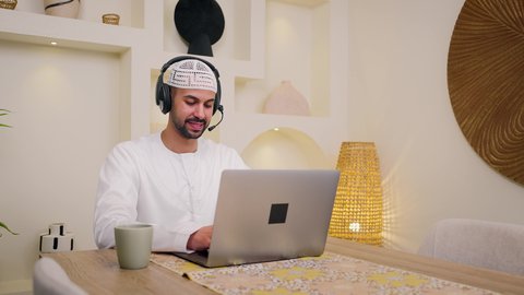 Listening to music through headphones, browsing apps and websites, quiet sessions at home, watching movies and series, integrating technology and modern techniques into daily life, holding an online business meeting, using a laptop, enjoying hot drinks, a close-up of an Emirati Gulf Arab man wearing a kandura sitting at home, a remote work session