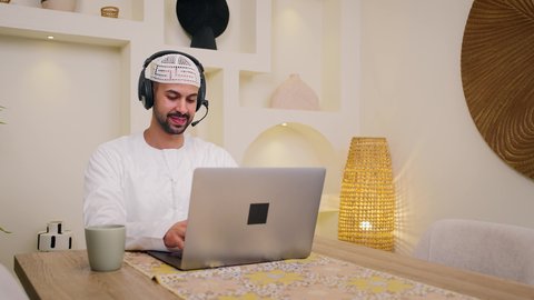 تصفح التطبيقات والمواقع ، جلسات هادئة في المنزل، مشاهدة الافلام والمسلسلات، دمج التكنولوجيا والتقنيات الحديثه فى الحياه اليوميه، عقد إجتماع عمل عبر الإنترنت، استخدام جهاز الحاسوب المحمول ،الاستماع الى الموسيقي عبر سماعات الاذن ، الاستمتاع بالمشروبات الساخنه ،تصوير مقرب لرجل عربي خليجي اماراتي يرتدي الكندورة يجلس في المنزل، جلسة عمل عن بعد