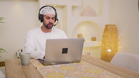 Listening to music through headphones, watching movies and series, integrating technology and modern techniques into daily life, holding an online business meeting, using a laptop, enjoying hot drinks, quiet sessions at home, a close-up of an Emirati Gulf Arab man wearing a kandura sitting at home, a remote work session, browsing applications and websites