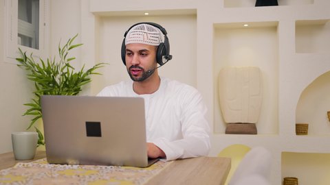 مشاهدة الافلام والمسلسلات، دمج التكنولوجيا والتقنيات الحديثه فى الحياه اليوميه، عقد إجتماع عمل عبر الإنترنت، استخدام جهاز الحاسوب المحمول ،الاستماع الى الموسيقي عبر سماعات الاذن ، الاستمتاع بالمشروبات الساخنه ، جلسات هادئة في المنزل، تصوير مقرب لرجل عربي خليجي اماراتي يرتدي الكندورة يجلس في المنزل، جلسة عمل عن بعد، تصفح التطبيقات والمواقع