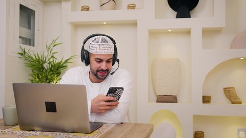 دمج التكنولوجيا والتقنيات الحديثه فى الحياه اليوميه، عقد إجتماع عمل عبر الإنترنت، استخدام جهاز الحاسوب المحمول والجوال ،الاستماع الى الموسيقي عبر سماعات الاذن ، الاستمتاع بالمشروبات الساخنه ، جلسات هادئة في المنزل، تصوير مقرب لرجل عربي خليجي اماراتي يرتدي الكندورة يجلس في المنزل، جلسة عمل عن بعد، تصفح التطبيقات والمواقع، مشاهدة الافلام والمسلسلات