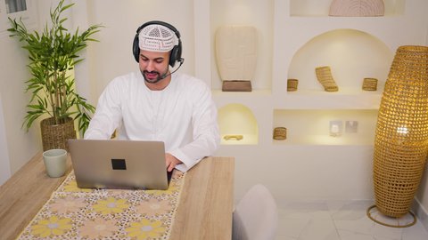 عقد إجتماع عمل عبر الإنترنت، استخدام جهاز الحاسوب المحمول ،الاستماع الى الموسيقي عبر سماعات الاذن ، الاستمتاع بالمشروبات الساخنه ، جلسات هادئة في المنزل، دمج التكنولوجيا والتقنيات الحديثه فى الحياه اليوميه، تصوير مقرب لرجل عربي خليجي اماراتي يرتدي الكندورة يجلس في المنزل، جلسة عمل عن بعد، تصفح التطبيقات والمواقع، مشاهدة الافلام والمسلسلات