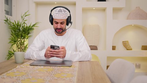 Using a mobile phone, listening to music through earphones, enjoying hot drinks, quiet sessions at home, integrating technology and modern techniques into daily life, a close-up of an Emirati Gulf Arab man wearing a kandura sitting at home, holding an online business meeting, a remote work session, browsing applications and websites, watching movies and series