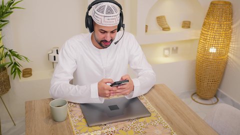 Listening to music through earphones, enjoying hot drinks, using a mobile phone, quiet sessions at home, integrating technology and modern techniques into daily life, a close-up of an Emirati Gulf Arab man wearing a kandura sitting at home, holding an online business meeting, a remote work session, browsing applications and websites, watching movies and series