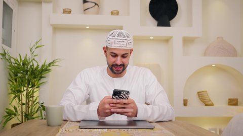 Enjoying hot drinks, using a mobile phone, having quiet sessions at home, incorporating modern technology into daily life, a close-up of an Emirati Gulf Arab man wearing a kandura sitting at home, holding an online business meeting, a remote work session, browsing apps and websites, watching movies and TV series.