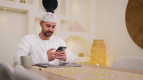 استخدام جهاز الهاتف المحمول ، جلسات هادئة في المنزل، دمج التكنولوجيا والتقنيات الحديثه فى الحياه اليوميه، تصوير مقرب لرجل عربي خليجي اماراتي يرتدي الكندورة يجلس في المنزل، عقد إجتماع عمل عبر الإنترنت، جلسة عمل عن بعد، تصفح التطبيقات والمواقع، الاستمتاع بالمشروبات الساخنه ، مشاهدة الافلام والمسلسلات