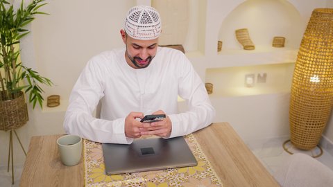 Quiet sessions at home, integrating technology and modern techniques into daily life. A close-up of an Emirati Gulf Arab man wearing a kandura sitting at home. Holding an online business meeting. Working remotely. Browsing apps and websites. Enjoying hot drinks, watching movies and TV series, using a mobile phone.
