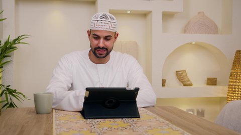 A close-up of an Emirati Gulf Arab man wearing a kandura and ghutra sitting at home, holding an online business meeting, working remotely, browsing apps and websites, enjoying a hot beverage, watching movies and TV series, having a quiet time at home, and using a tablet.