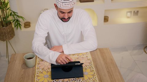 A remote working session, browsing apps and websites, enjoying a hot beverage, watching movies and TV series, holding an online business meeting, a close-up of an Emirati Gulf Arab man wearing a kandura and ghutra sitting at home, quiet sessions at home, using a tablet.
