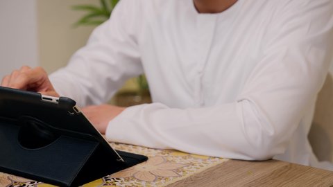 تصفح التطبيقات والمواقع، الاستمتاع بالمشروبات الساخنه ، مشاهدة الافلام والمسلسلات ،عقد إجتماع عمل عبر الإنترنت، تصوير مقرب لرجل عربي خليجي اماراتي يرتدي الكندورة والغتره يجلس في المنزل، جلسة عمل عن بعد، جلسات هادئة في المنزل، استخدام جهاز التابلت