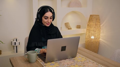 عقد إجتماع عمل عبر الإنترنت،  جلسات هادئة في المنزل، مشاهدة الافلام والمسلسلات ، تصفح التطبيقات والمواقع، الاستمتاع بالمشروبات الساخنه ، الاستماع الى الموسيقي عبر سماعات الاذن ،تصوير مقرب لامرأة عربية خليجية سعودية ترتدي العباءه والحجاب تجلس في المنزل، جلسة عمل عن بعد، سيدة اماراتيه تستخدم جهاز الحاسوب المحمول