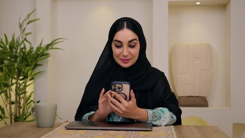 تصوير مقرب لامرأة عربية خليجية سعودية ترتدي العباءه والحجاب تجلس في المنزل، اجراء مكالمات الفيديو ، تصفح التطبيقات والمواقع عبر الجوال ، جلسات هادئة في المنزل،دمج التقنيات الحديثه فى الحياه اليوميه ، الاستماع الى الموسيقي عبر سماعات الاذن ، سيدة اماراتيه تستخدم جهاز الهاتف المحمول  ، الاستمتاع بالمشروبات الساخنه