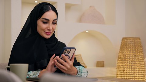 اجراء مكالمات الفيديو ، تصفح التطبيقات والمواقع عبر الجوال ، جلسات هادئة في المنزل،دمج التقنيات الحديثه فى الحياه اليوميه ، الاستماع الى الموسيقي عبر سماعات الاذن ،تصوير مقرب لامرأة عربية خليجية سعودية ترتدي العباءه والحجاب تجلس في المنزل، سيدة اماراتيه تستخدم جهاز الهاتف المحمول  ، الاستمتاع بالمشروبات الساخنه