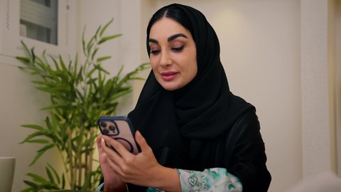 تصفح التطبيقات والمواقع عبر الجوال ، جلسات هادئة في المنزل،دمج التقنيات الحديثه فى الحياه اليوميه ، مشاهدة الافلام والمسلسلات ، الاستماع الى الموسيقي عبر سماعات الاذن ،تصوير مقرب لامرأة عربية خليجية سعودية ترتدي العباءه والحجاب تجلس في المنزل، سيدة اماراتيه تستخدم جهاز الهاتف المحمول  ، الاستمتاع بالمشروبات الساخنه