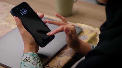 Integrating modern technologies into daily life, enjoying hot drinks, quiet sessions at home, watching movies and series, browsing applications and websites via mobile, listening to music through earphones, a close-up of a Saudi Arabian Gulf woman wearing an abaya and hijab sitting at home, an Emirati woman using a mobile phone