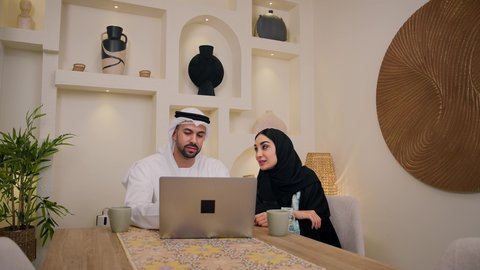 Holding an online business meeting, enjoying hot drinks, integrating technology and modern techniques into daily life, listening to music through earphones, gestures of concentration and integration, making video calls over the internet, an Arab Gulf Emirati man holding a laptop sitting at home with his wife, a veiled Arab Gulf Emirati woman using a tablet