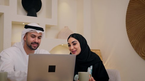 الاستمتاع بالمشروبات الساخنه، دمج التكنولوجيا والتقنيات الحديثه فى الحياه اليوميه، الاستماع الى الموسيقي عبر سماعات الاذن ، إيماءات التركيز والاندماج، إجراء مكالمات فيديو عبر الانترنت، رجل عربي خليجي اماراتي يمسك جهاز الحاسوب المحمول يجلس فى المنزل مع زوجته، عقد إجتماع عمل عبر الإنترنت، إمرأة عربية خليجية اماراتيه محجبة تستخدم جهاز التابلت