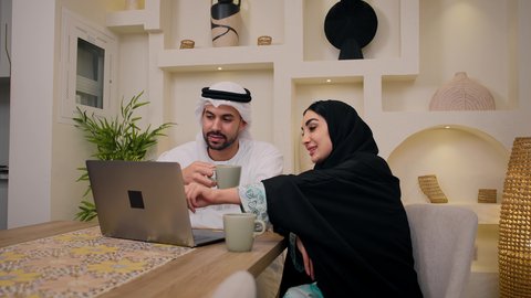 دمج التكنولوجيا والتقنيات الحديثه فى الحياه اليوميه، الاستماع الى الموسيقي عبر سماعات الاذن ، إيماءات التركيز والاندماج، الاستمتاع بالمشروبات الساخنه،  إجراء مكالمات فيديو عبر الانترنت، رجل عربي خليجي اماراتي يمسك جهاز الحاسوب المحمول يجلس فى المنزل مع زوجته، عقد إجتماع عمل عبر الإنترنت، إمرأة عربية خليجية اماراتيه محجبة تستخدم جهاز التابلت