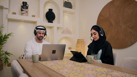 إيماءات التركيز والاندماج، الاستمتاع بالمشروبات الساخنه،  إجراء مكالمات فيديو عبر الانترنت، رجل عربي خليجي اماراتي يمسك جهاز الحاسوب المحمول يجلس فى المنزل مع زوجته، دمج التكنولوجيا والتقنيات الحديثه فى الحياه اليوميه، عقد إجتماع عمل عبر الإنترنت، إمرأة عربية خليجية اماراتيه محجبة تستخدم جهاز التابلت ، الاستماع الى الموسيقي عبر سماعات الاذن
