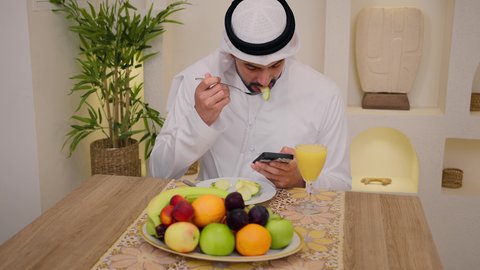 Quiet morning atmosphere, close-up of an Emirati Gulf Arab man wearing a kandura and ghutra, sitting at a table at home, using a mobile phone while eating a healthy breakfast, fresh fruit slices and orange juice, a healthy lifestyle, integrating modern technology into daily routine.