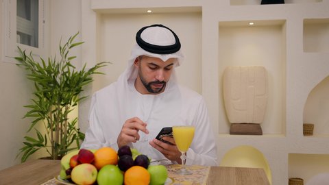 Healthy lifestyle, calm morning atmosphere, close-up of an Emirati Gulf Arab man wearing a kandura and ghutra, sitting at a table at home, using a mobile phone while eating a healthy breakfast, fresh fruit slices and orange juice, integrating modern technology into daily routine.