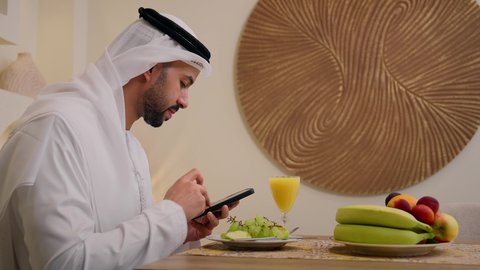 Fresh fruit slices and orange juice, a healthy lifestyle, a calm morning atmosphere, a close-up of an Emirati Gulf Arab man wearing a kandura and ghutra, sitting at a table at home, using a mobile phone while eating a healthy breakfast, integrating modern technology into his daily routine.