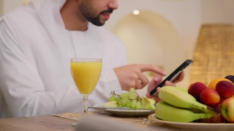 Eating a healthy breakfast, integrating modern technology into daily routine, fresh fruit slices and orange juice, a healthy lifestyle, a calm morning atmosphere, a close-up of an Arab Gulf Emirati man wearing a kandura and ghutra, sitting at a table at home using a mobile phone.