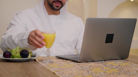 Close-up shot of an Emirati Gulf Arab man wearing a kandura and ghutra, sitting at a table at home using a mobile phone, eating a healthy breakfast, integrating modern technology into daily routine, fresh fruit slices and orange juice, a healthy lifestyle, and a calm morning atmosphere.