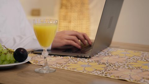 الاستمتاع بشرب المشروبات الباردة ، تصوير مقرب لرجل عربي خليجي إماراتي يرتدي الكندورة والغترة، يجلس على طاولة في المنزل ويستخدم الهاتف المحمول ،تناول وجبة فطور صحية، دمج التكنولوجيا الحديثة مع الروتين اليومي ، شرائح الفاكهة الطازجة وعصير البرتقال، نمط حياة صحي، أجواء صباحية هادئة