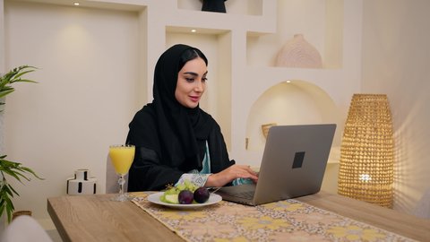 دمج التكنولوجيا الحديثة مع الروتين اليومي ، شرائح الفاكهة الطازجة وعصير البرتقال، الاستمتاع بشرب المشروبات الباردة ، نمط حياة صحي، تصوير مقرب لامراه عربية خليجية سعودية ترتدي العباءة والحجاب، تجلس على طاولة في المنزل ويستخدم الهاتف المحمول ،سيدة اماراتية تستمتع بتناول وجبة فطور صحية، أجواء صباحية هادئة