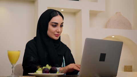 Enjoying cold drinks, fresh fruit slices and orange juice, integrating modern technology with daily routine, healthy lifestyle, close-up of a Saudi Arabian Gulf woman wearing abaya and hijab, sitting at a table at home using a mobile phone, Emirati lady enjoying a healthy breakfast, calm morning atmosphere