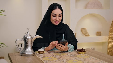 دمج التكنولوجيا الحديثة مع الروتين اليومي ،سيدة عربية خليجية إماراتية ترتدي العباءة والحجاب، تقوم بصب القهوة العربية في فنجان، أجواء منزلية دافئة ، طبق تمر سعودي ، امراه سعودية تستخدم الهاتف المحمول ، جلسة ضيافة تقليدية.