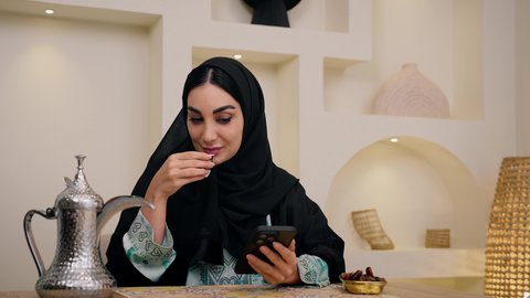 طبق تمر سعودي ، دمج التكنولوجيا الحديثة مع الروتين اليومي ،سيدة عربية خليجية إماراتية ترتدي العباءة والحجاب، تقوم بصب القهوة العربية في فنجان، امراه سعودية تستخدم الهاتف المحمول ، جلسة ضيافة تقليدية ، أجواء منزلية دافئة