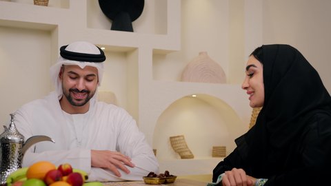 رجل اماراتي يرتدي الكندورة والغترة ويقوم بصب القهوة العربية في الفنجان لزوجته، لحظات ودية مليئة بالاحترام والمحبة، دمج العادات والتقاليد مع دفء الحياة اليومية، أجواء ضيافة عربية أصيلة تعكس التراث الخليجي، زوجان عربيان خليجيان اماراتيان يجلسان معًا في أجواء منزلية دافئة